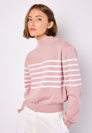 Maglione rosa con un collo alto a coste, presentante strisce orizzontali bianche, texture morbida e vestibilità rilassata. Abbinato a pantaloni di colore chiaro.