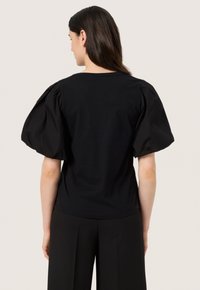 Sort kortærmet top med puffede skuldre, rund halsudskæring og et glat, let stof. Bagfra vises rene linjer og et minimalistisk design.