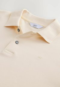 Crèmekleurige polo met een gestructureerde brei, twee donkere knopen en een geborduurd kroonlogo op de linkerborst. "NEXT" label zichtbaar aan de binnenkant van de kraag.