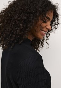 Svart stickad tröja med ribbat mönster på axlarna, hög halsringning och långa ärmar. Modellen ler och ses från sidan.