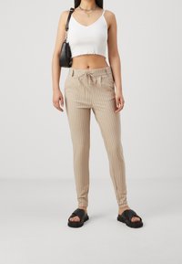 Pantalons rayés beiges avec une taille à cordon, associés à un crop top blanc. Le mannequin porte des sandales noires à enfiler et porte un petit sac noir.