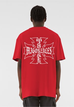 Punainen puuvillainen t-paita, jossa on suuri valkoinen risti selässä ja tyyliteltyyn fonttiin kirjoitettu teksti "MIGONZALES". Lyhyet hihat.