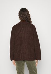 Personne aux longs cheveux bouclés vue de dos, portant un pull en maille épaisse marron foncé et un pantalon en velours côtelé vert.