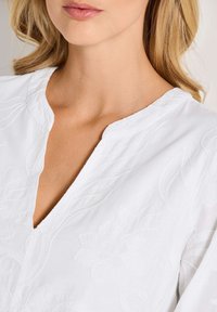 Nahaufnahme des Halses und des oberen Brustbereichs einer Frau, die eine weiße Bluse mit zarten Blumenstickereien und einem gerundeten V-Ausschnitt trägt.