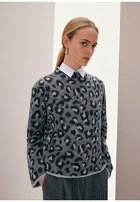 Vrouw met blond haar in een paardenstaart, draagt gouden oorbellen, een grijze trui met luipaardprint over een witte blouse en een grijze streepbroek.