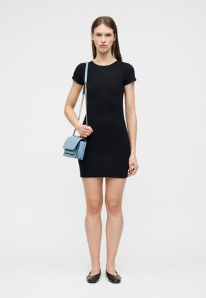 Kvinne står iført en tettsittende svart kjole med korte ermer, svarte flate sko, og holder en liten lyst blå skulderveske med en gullkjede.