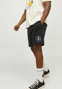Pantaloni corti neri con logo di testo bianco sulla gamba sinistra, indossati con una t-shirt grafica beige chiaro e scarpe nere. Il tessuto appare morbido e informale.