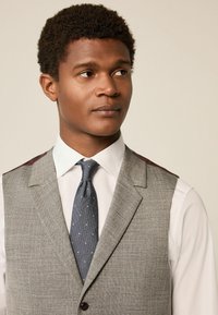 Pelēka houndstooth veste ar bordo akcentiem, apvienota ar baltu krekla un tumši zilu kaklasaiti, kas rotāta ar baltām punktiem. Gluda tekstūra, pielāgota piegriezuma.
