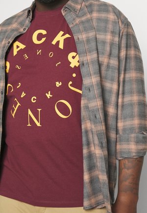 Man draagt een bordeauxrood t-shirt met verspreide gele letters en een grijs-perzikkleurig geruit overhemd erover, mouw opgerold.
