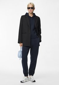 Blazer nero sopra un hoodie blu navy, abbinato a joggers blu navy e sneakers bianche. L'outfit include una borsa azzurro chiaro e occhiali da sole scuri.