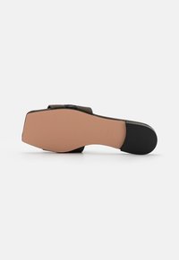 Sandal avec un bout carré et une semelle plate, fond tan avec un patch de talon noir, exposé semelle vers le haut sur un fond blanc.