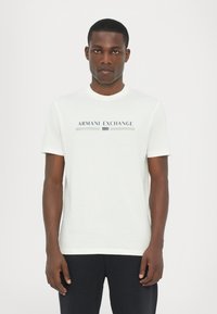 Fehér, rövid ujjú póló kerek nyakkivágással, amelyen kék és zöld színű "ARMANI EXCHANGE 1991" grafika található. Puha anyag, sima textúrával.