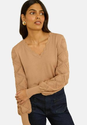 Ljusbrun stickad tröja med en vågmönstrad halsringning, texturerade mönstrade ärmar och en ledig passform, kombinerad med mörkblå jeans.