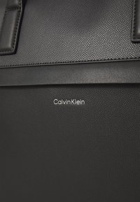 Schwarze, strukturiert Leder Tasche mit einem glatten Riemen und geprägtem Calvin Klein Logo. Die Oberfläche ist mit einer gepunkteten Optik verziert.