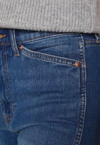 Tmavě modré džíny z denimu s těsným střihem, které mají měděné kování na přední kapse a oranžové prošívání podél švů.