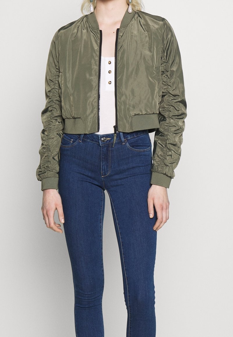 Femme portant une veste bomber courte vert olive, un haut blanc boutonné et un jean skinny bleu foncé sur un fond clair uni.