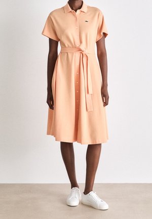 Vrouw draagt een perzikkleurige midi-jurk met knopen, een tailleriem die is gestrikt en witte sneakers, staand tegen een effen lichte achtergrond.