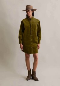 Mulher com um vestido de corduroy botão em verde oliva, botas castanhas até o tornozelo e um chapéu de abas largas em tom bege com uma fita para o queixo, de pé contra um fundo simples.