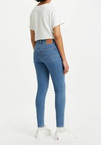 Jeans ajustados de mezclilla azul claro con cintura alta, que cuentan con bolsillos traseros y una etiqueta de cuero en la cinturilla, combinados con una camiseta blanca.