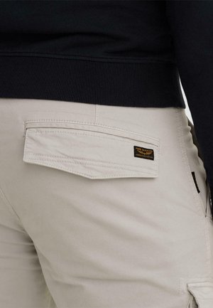 Pogled od zadaj na bež hlače z žepom s poklopcem, na katerem je oznaka »PME Legend American Classic«, nošene z črnim rebrastim puloverjem.