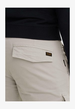 Zadný pohľad na béžové nohavice s klopovou vreckovou, na ktorej je štítok "PME Legend American Classic", nosené s čiernym rebrovaným svetrom.