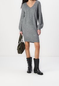 Szary dzianinowy sweter midi z dekoltem w serek, z długimi bufiastymi rękawami, sięgający do kolan. Stylizowany z teksturowaną oliwkową torebką i czarnymi wysokimi botkami z guzikami.