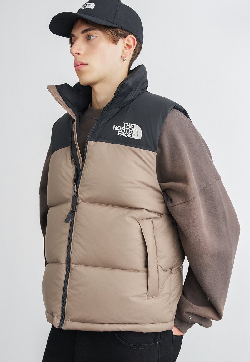 Beige puffer veste ar melnu augšējo daļu. Iezīmē stāvošu apkakli, rāvējslēdzēja aizdari, sānu kabatas un logotipu uz krūtīm.