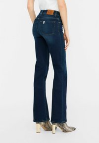 Jeans acampanados de color azul oscuro con cintura alta, que presentan un parche de cuero marrón claro y bolsillos traseros, combinados con tacones de serpiente con detalles en plata.