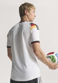 Homme blond en maillot de football blanc avec des rayures noires, rouges et jaunes tient un ballon de football multicolore, de profil sur un fond uni.