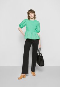 Blusa smock em verde menta com mangas bufantes, bainha peplum, combinada com jeans pretos de boca de sino e mules beges de ponta aberta, segurando uma bolsa preta.