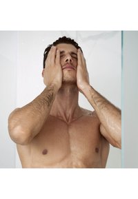 Un uomo a petto nudo si trova sotto la doccia, con gocce d'acqua sulla pelle, le mani sul viso, di fronte a una parete doccia in vetro trasparente.
