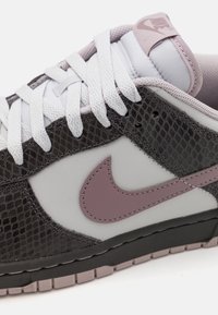 Nike Dunk Low in grijs en zwart met een textuur in slangenprint, witte veters en een mauve swoosh. Rubberen zool met roze accenten.