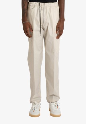 Persona che indossa pantaloni beige con coulisse e sneakers bianche con suola in gomma, in piedi davanti a uno sfondo chiaro uniforme.