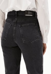 Närbild av en person som bär hög midja svarta jeans med bakfickor och en vit skjorta instoppad, där varumärkesetiketten på midjebandet syns.