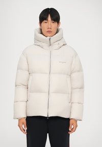 Puffjacka i ljusbeige med hög krage, dragkedjestängning, stora vadderade sektioner och minimal logotyp på framsidan.