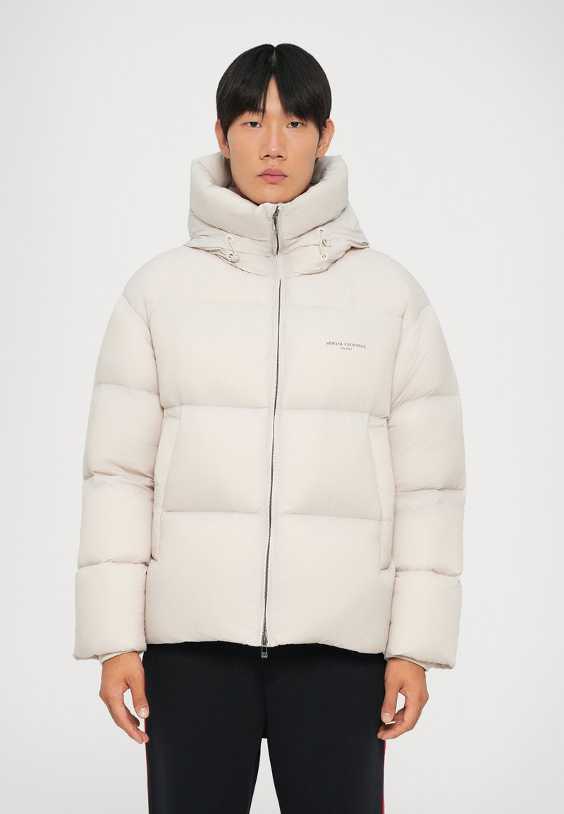Puff jakke i lys beige med høj krave, lynlås lukning, store quiltede sektioner og minimal branding på forsiden.