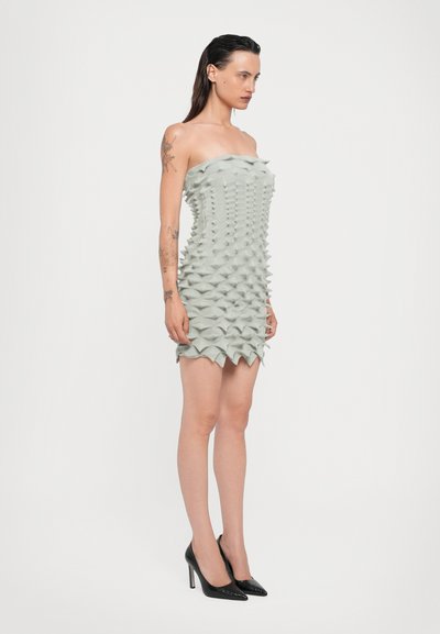 Vestido sin tirantes en verde claro, con un diseño texturizado en 3D de picos, silueta ajustada y dobladillo con volantes. Combinado con tacones altos negros.