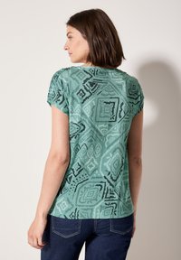 Chemise à manches courtes vert à motif géométrique noir, avec une coupe décontractée et un léger froncé aux épaules.