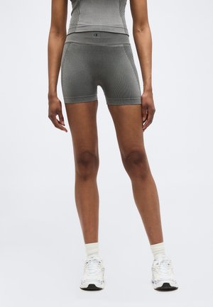 Persona che indossa pantaloncini grigi aderenti a vita alta e top coordinato, in piedi dritta, con calzini bianchi e scarpe sportive.