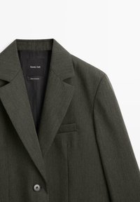 Blazer su misura verde scuro con colletto a cappotto, chiusura a un bottone e taschino sul petto. Texture liscia e motivo sottile. Etichetta del marchio visibile.