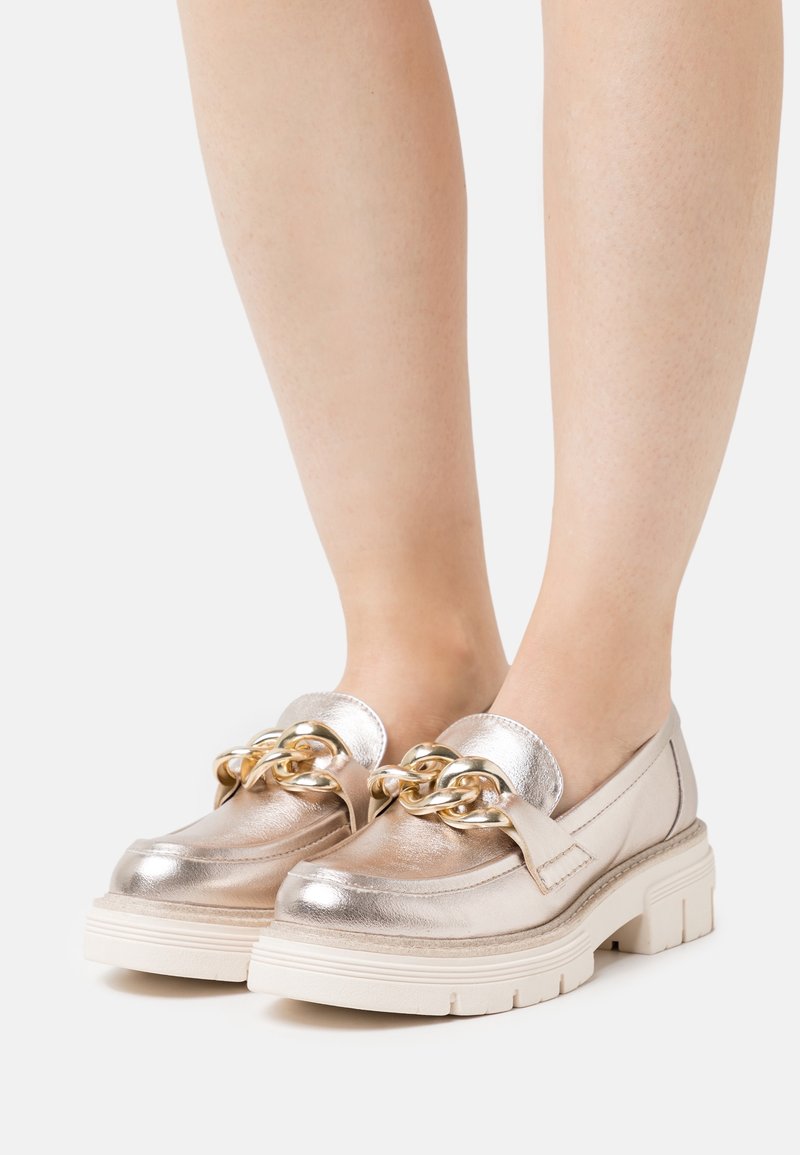 Metallisch goldene Loafers mit einer klobigen weißen Sohle, die ein dekoratives Goldketten-Detail im oberen Bereich aufweisen. Glatte Textur und runde Form.