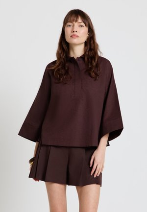 Frau mit langen braunen Haaren, die eine lockere dunkelbraune Bluse mit weiten Ärmeln und passende plissierte Shorts vor einfarbigem Hintergrund trägt.