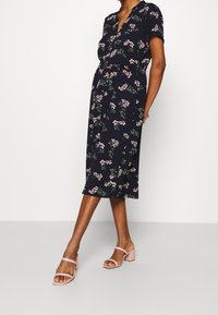 Donna che indossa un vestito midi nero con stampa floreale rosa e maniche corte, abbinato a sandali con tacco blocco rosa chiaro, su uno sfondo neutro.