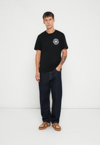 Svart bomull T-skjorte med rund hals og hvitt grafisk trykk. Kombinert med løstsittende mørk denimjeans og brune og beige joggesko.