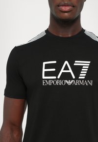 Juodas trumpomis rankovėmis marškinėliai su balta "EA7 Emporio Armani" logotipu, turintys dryžuotus peties akcentus ir prigludusią dizaino formą.