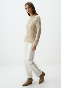 Beige gestrickter Pullover mit strukturiertem Muster, kombiniert mit weißen Hosen und hellbraunen westerninspirierten Stiefeletten mit dekorativer Stickerei.