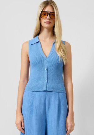 Femme blonde portant des lunettes de soleil oversize teintées orange, un top en maille bleu clair sans manches à boutons et un pantalon ample texturé assorti.