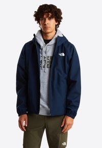 Veste imperméable bleu marine avec une capuche, dotée de poignets élastiques et d'un logo sur la poitrine, portée sur un sweat à capuche gris et un pantalon olive.