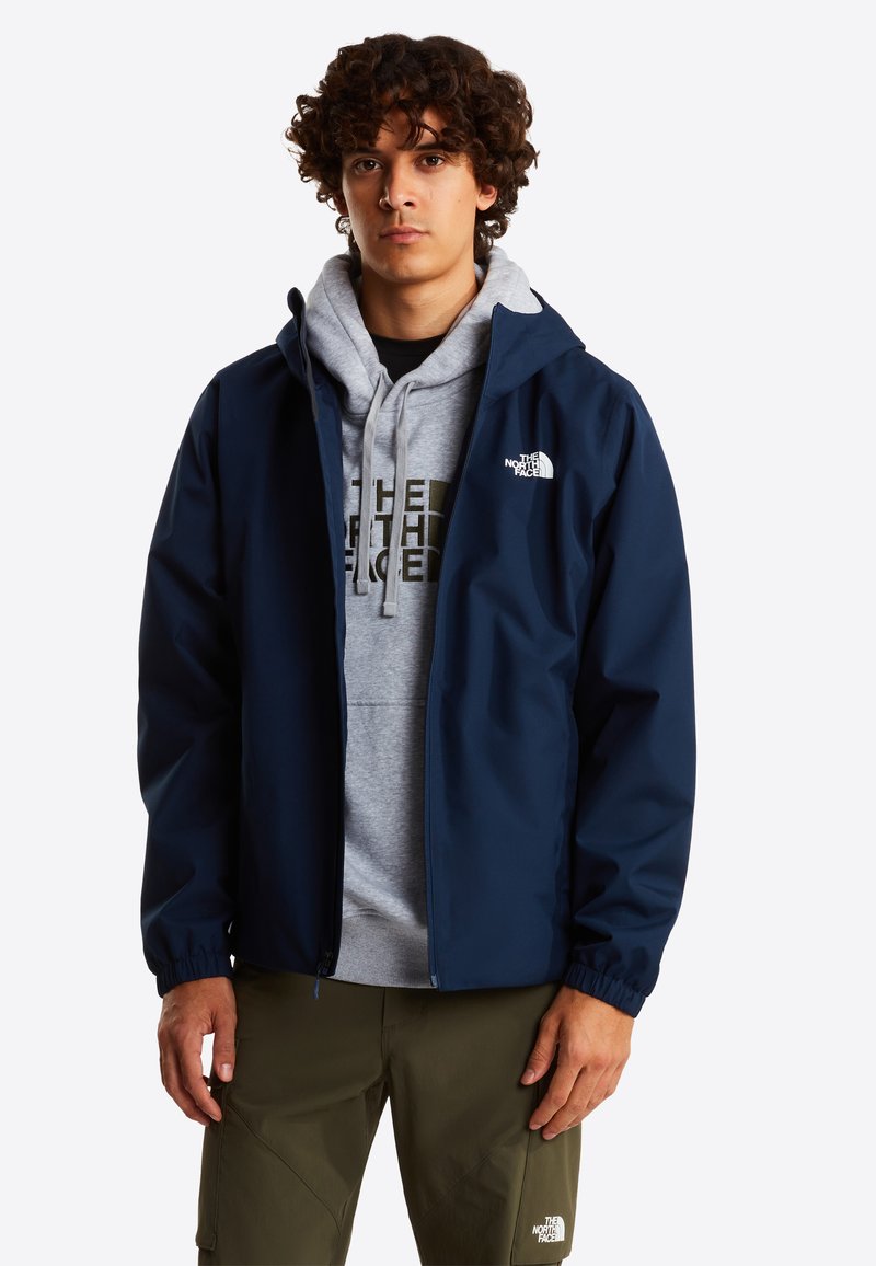 Veste imperméable bleu marine avec une capuche, dotée de poignets élastiques et d'un logo sur la poitrine, portée sur un sweat à capuche gris et un pantalon olive.