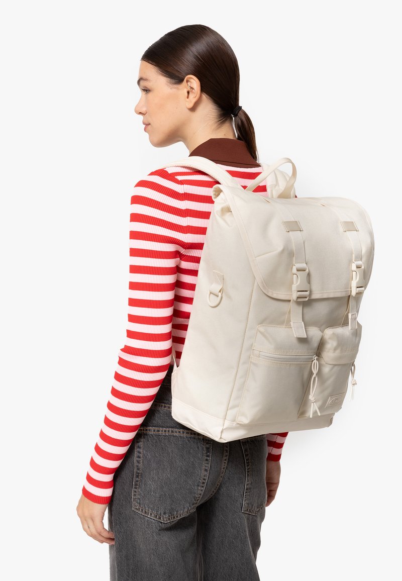 Zaino in tela beige con spallacci regolabili, due tasche frontali e dettagli in metallo. Il modello indossa una maglietta a righe rosse e bianche.